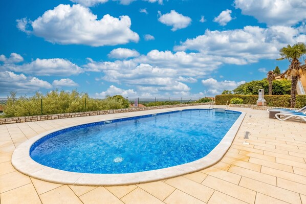 A swimming pool with a tiled deck, surrounded by greenery and palm trees, under a clear sky.