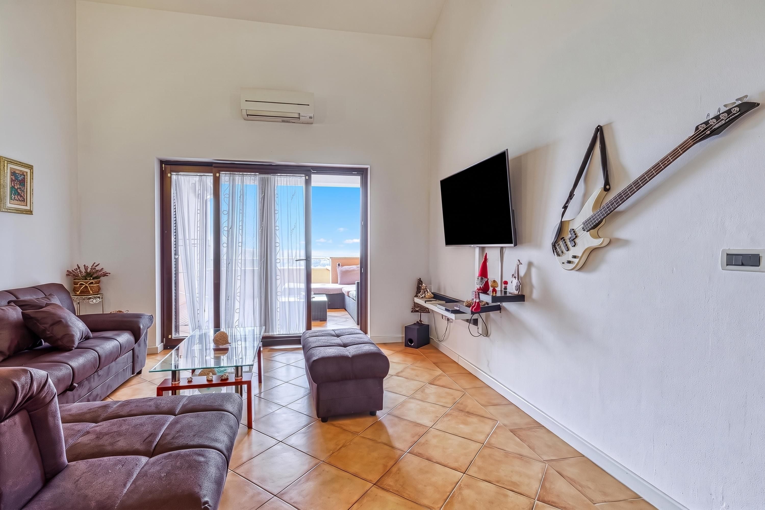A living room with a sofa, a glass coffee table, a TV mounted on the wall, and a sliding glass door leading to an outdoor area.