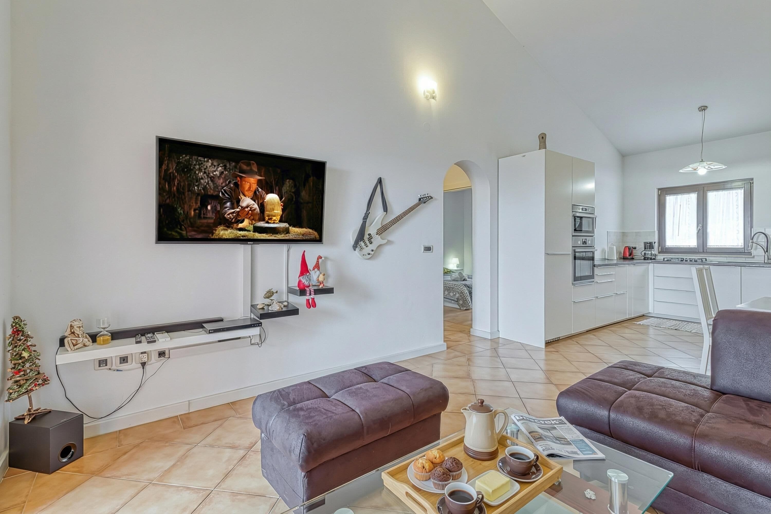 Een moderne woonkamer met een flatscreen televisie, een bank, een bijzettafeltje met een schaal eten, en een keuken op de achtergrond.