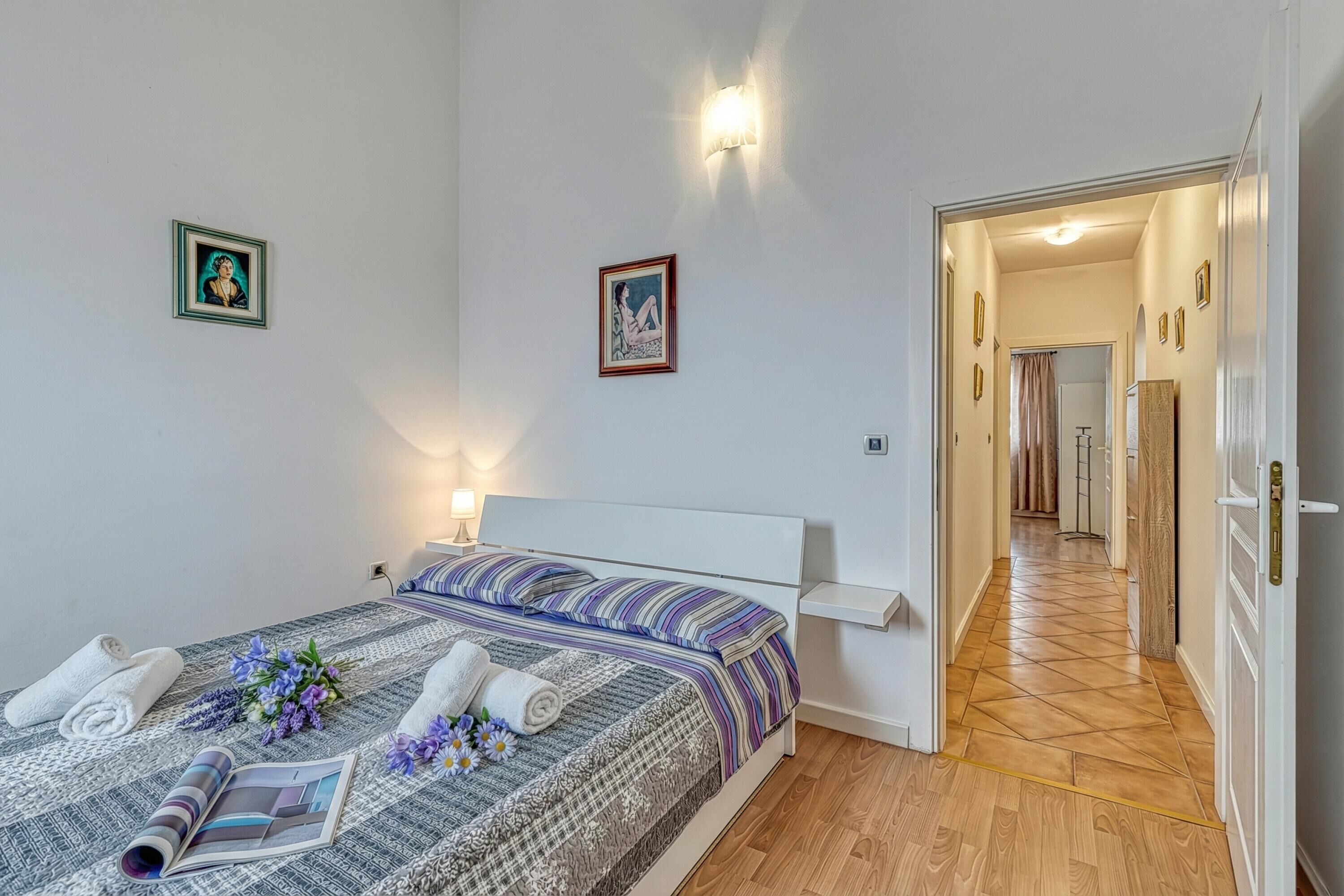 A bedroom with a bed, bedside tables, wall-mounted light fixtures, and a hallway leading to another room.