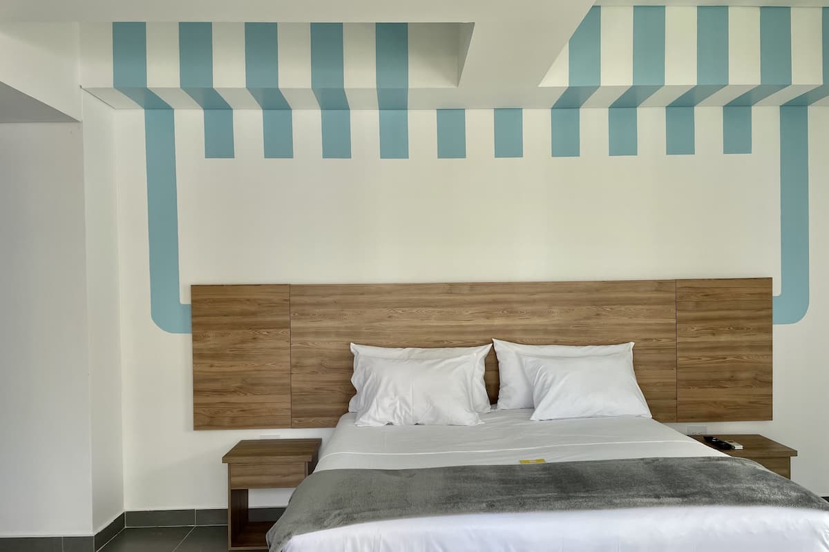 Un lit bien fait, avec une tête de lit en bois, des oreillers blancs et une couverture grise. Une table de chevet en bois se trouve à côté du lit, sur laquelle repose une télécommande. La pièce présente un design moderne, avec un mur de couleur claire et un plafond à motifs.