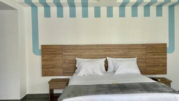 Una cama bien hecha con cabecera de madera, almohadas blancas y una manta gris. Junto a la cama hay una mesita de noche con un control remoto. La habitación tiene un diseño moderno con una pared de color claro y un techo con diseño.