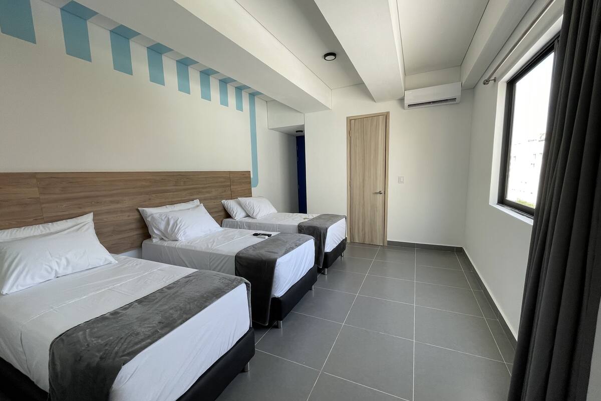 Une chambre d’hôtel avec trois lits, une tête de lit en bois, une grande fenêtre avec des rideaux et un sol carrelé.