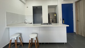 Una cocina moderna con encimera blanca, dos taburetes, un refrigerador de acero inoxidable y una puerta azul al fondo.