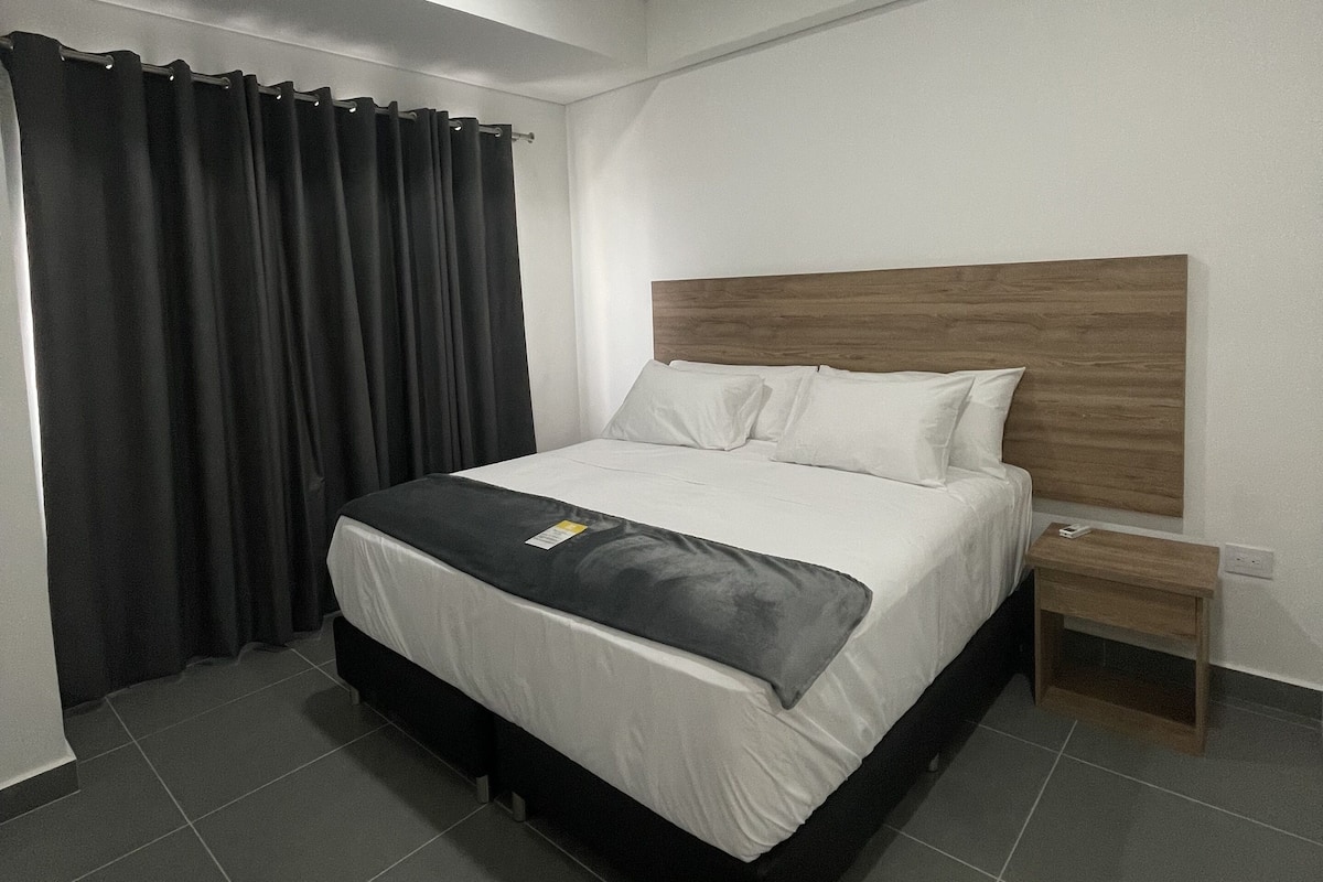 Une chambre d’hôtel moderne avec un grand lit, une tête de lit en bois, une table de chevet et un rideau.