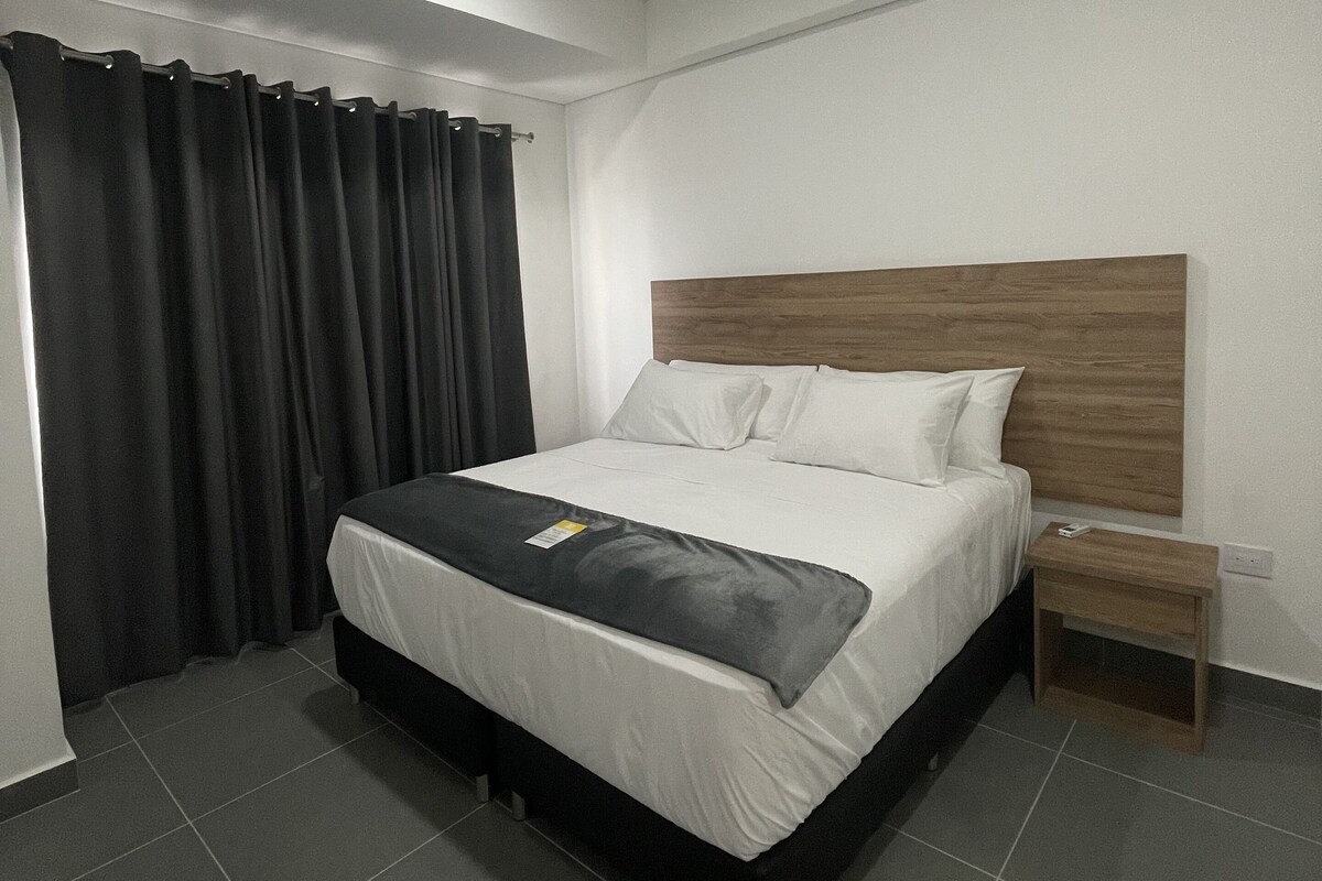 A modern hotel room with a large bed, a wooden headboard, a bedside table, and a curtain.