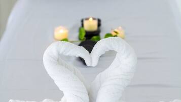 A bed with white linens, a heart-shaped towel arrangement, and candles on the nightstand.