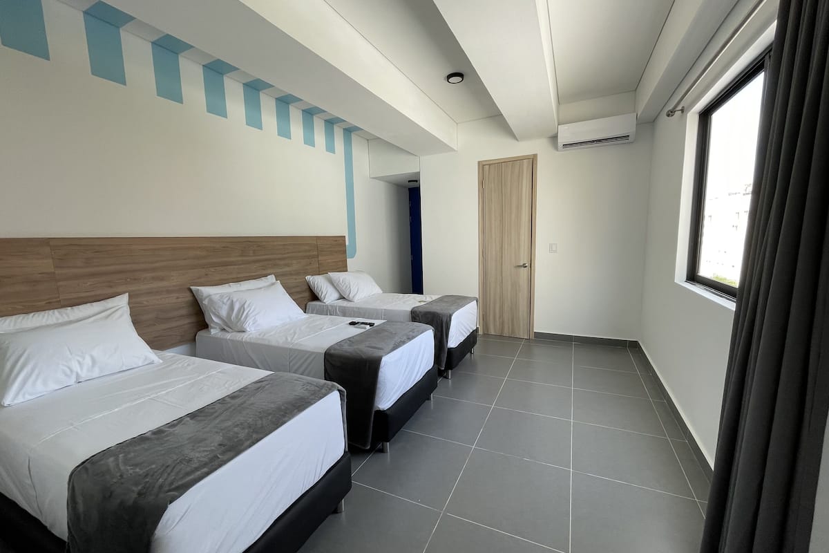 Une chambre d’hôtel avec trois lits, une tête de lit en bois, une grande fenêtre et un sol carrelé.