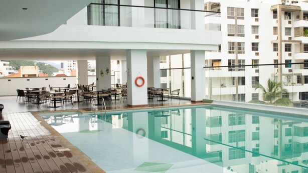 Un área de piscina en la azotea con barandilla de cristal, mobiliario exterior y vistas de edificios circundantes.