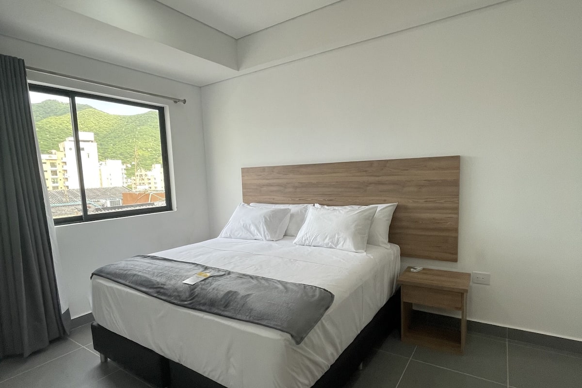 Une chambre moderne avec un grand lit, une tête de lit en bois, une table de chevet et une fenêtre donnant sur des bâtiments et des collines.