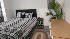 A bedroom with a large bed, a small round table, a wooden nightstand, a potted plant, and a window.
