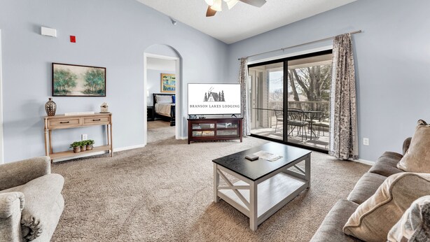 A living room with a ceiling fan, a large window with curtains, a sofa, a coffee table, and a TV displaying 'Branson Lakes Lodging'. - Gorgeous, Pools, Hot Tub, Golf, 3 Min To Strip! (Branson)