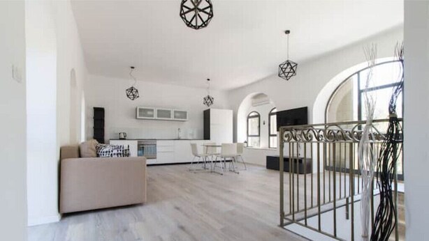 Amplia sala de estar con sofá, mesa de comedor y sillas. Se observa una cocina al fondo con electrodomésticos y armarios modernos. El ambiente tiene un techo alto con lámparas decorativas.