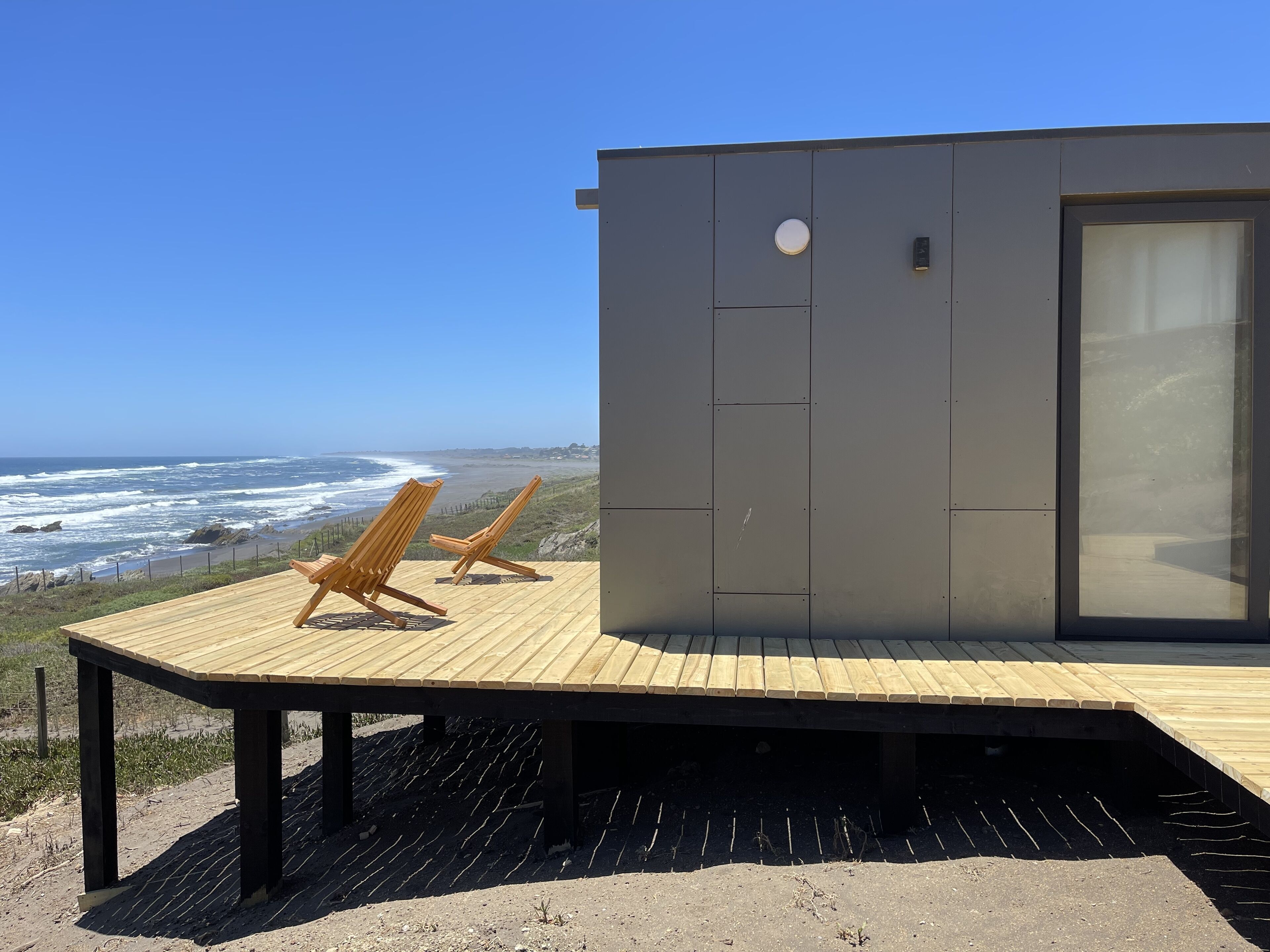 Una casa moderna frente al mar, con terraza de madera, dos sillones de descanso y un amplio ventanal con vistas al océano.