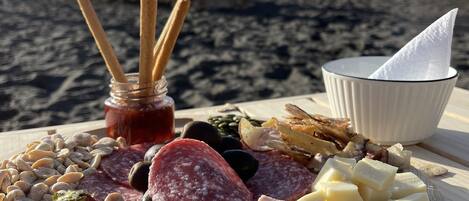 Una tabla de madera con diversos quesos, carnes y frutas en una playa, con sombrillas al fondo.