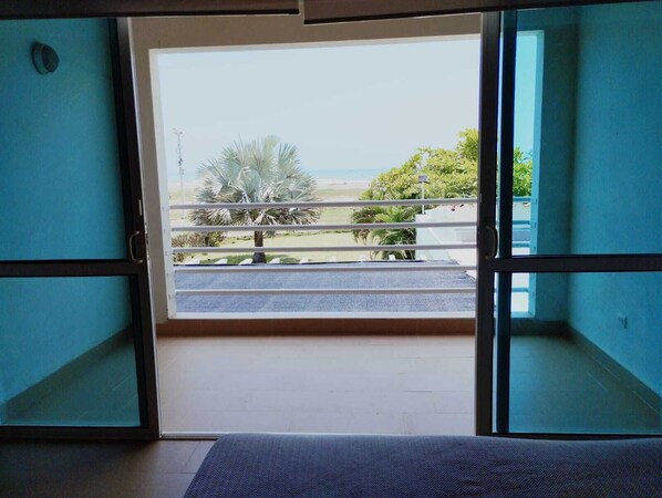 A room with a sliding glass door leading to a balcony with a view of the beach and palm trees.
