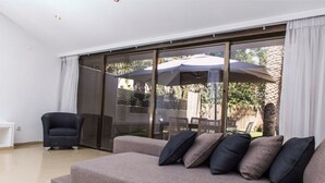 Ein modernes Wohnzimmer mit grauem Sofa, schwarzem Sessel und einer Glasschiebetür, die auf eine Terrasse mit Gartenmöbeln und Sonnenschirmen führt.