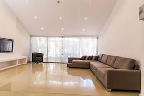 A spacious living room with a large brown sofa, a small armchair, and a flat-screen TV mounted on the wall.
