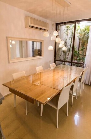 A modern dining room with a large wooden table, white chairs, pendant lights, a large mirror, and a view of greenery through a window.