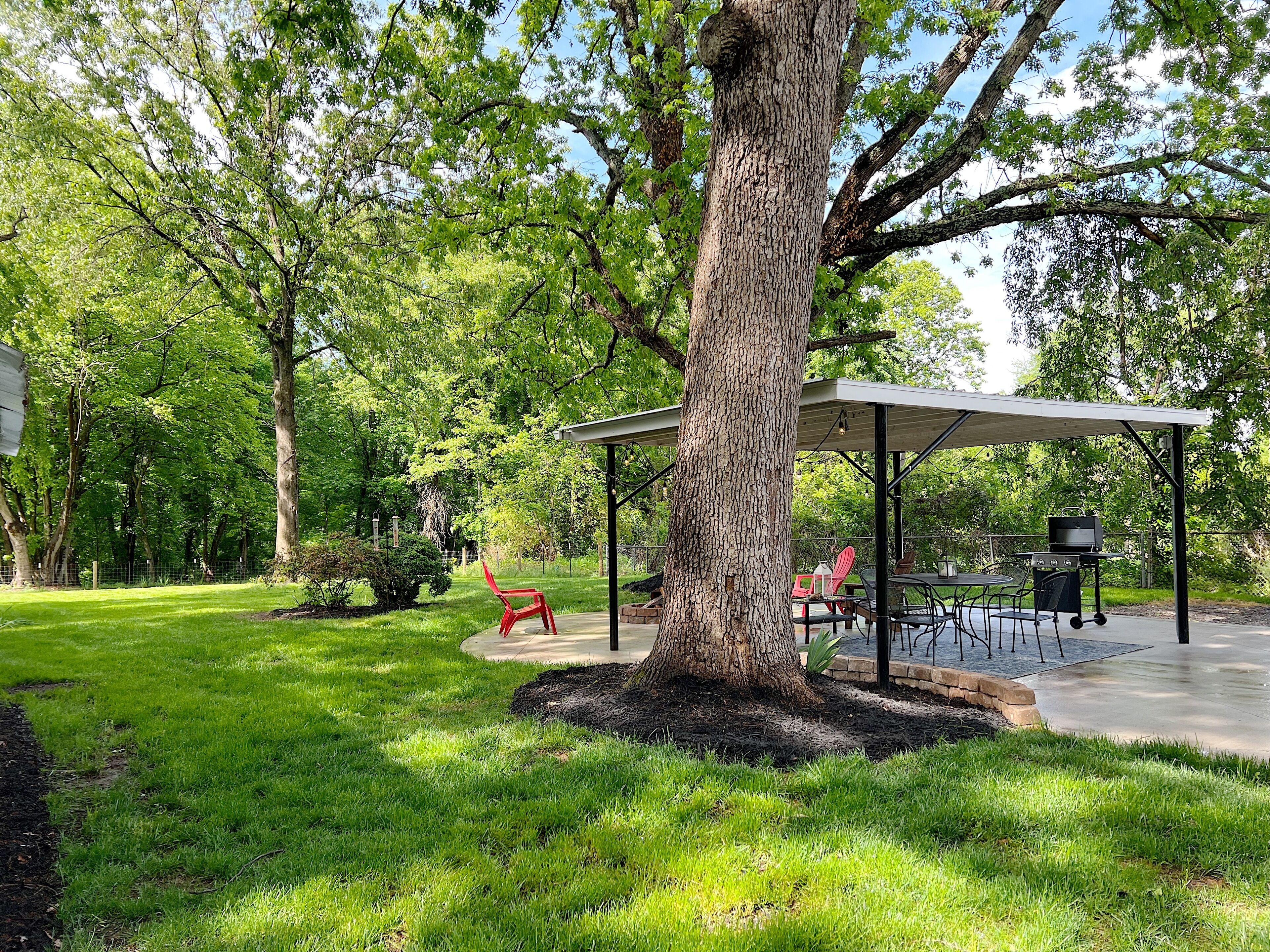 A covered outdoor seating area with a barbecue grill, surrounded by lush greenery and trees.