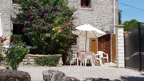 Un edificio in pietra con tetto in tegole, un piccolo giardino con piante fiorite, posti a sedere all'aperto e un vialetto sterrato.