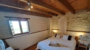 Una camera da letto con soffitto in legno, un letto grande con lenzuola bianche, un comodino con una lampada e una finestra con tende.