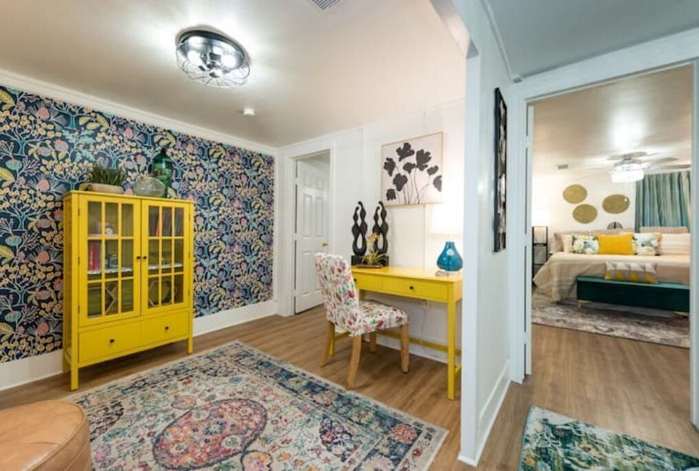 Una habitación con un papel tapiz de motivos florales, un armario amarillo, un escritorio, una silla y una cama con cabecera verde.
