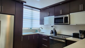 A modern kitchen with dark cabinets, a stainless steel microwave, an oven, and a window with blinds. - Aquatika Happy Place *pools and beach access* (Medianía Alta)