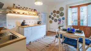 A modern kitchen with white cabinets, a wooden countertop, and a hanging light fixture. There is a dining area with a table set for two, blue chairs, and a vase with flowers. - Dione terrace double flat by Dimore in Sicily (Syracuse)