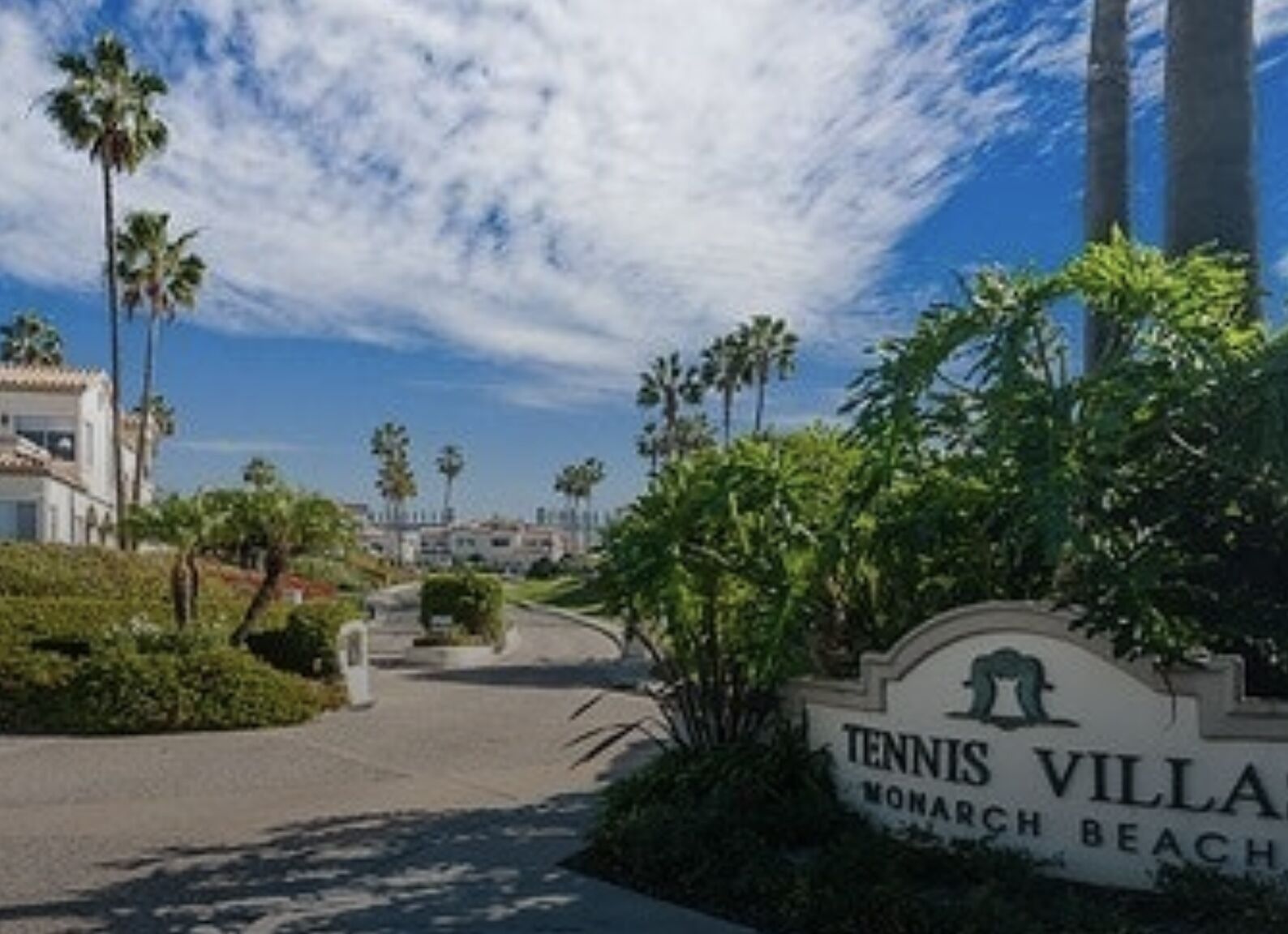 A sign for Tennis Village with a view of a palm tree-lined street and buildings in the background.