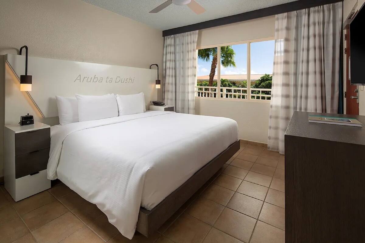 Habitación de hotel con cama, mesitas de noche, escritorio y vista a palmeras a través de la ventana.