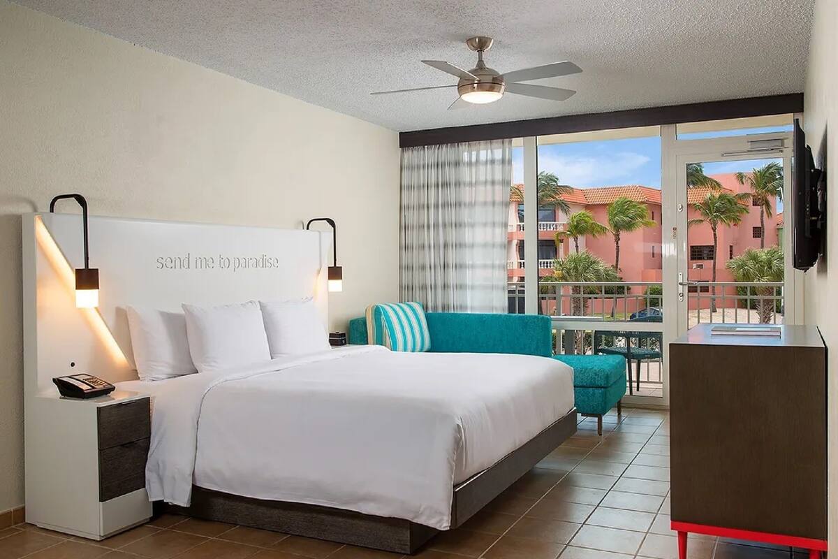 Una habitación de hotel con cama, mesita de noche con teléfono, una silla color turquesa, una pequeña mesa y un balcón con vistas a palmeras y edificios.