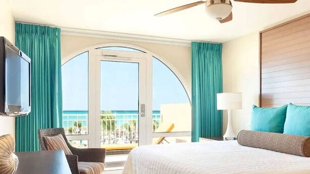 Une chambre d’hôtel équipée d’un lit, d’un bureau, d’une chaise, d’une télévision, d’un ventilateur de plafond et d’un balcon avec vue sur l’océan.