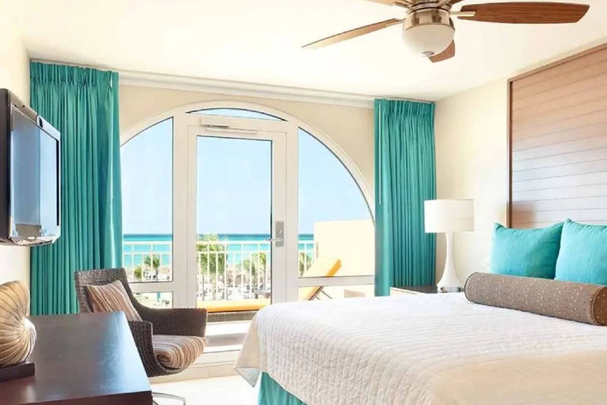 Habitación de hotel con cama, escritorio, silla, televisión, ventilador de techo y balcón con vista al océano.