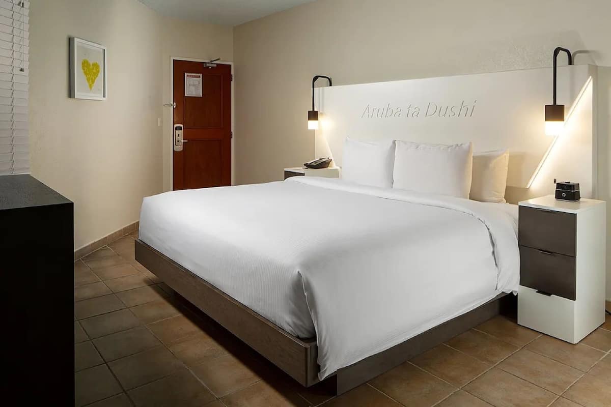 Una cama bien hecha con una colcha y almohadas blancas, una mesita de noche con una lámpara, y un cabecero fijado a la pared con el texto "Aruba Ta Dushi".