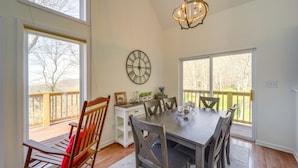 A dining area with a wooden table and chairs, a chandelier, a clock, and large windows overlooking a deck.