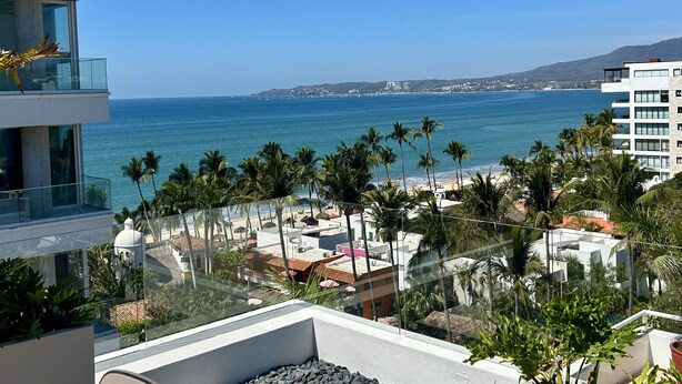 A coastal view from a balcony with a clear view of the ocean, palm trees, and buildings in the background. - Modern 2BR Golden Zone | Top Floor | Ocean View (Bucerias)