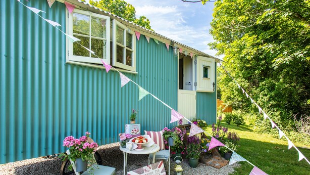 A small, single-story house with a corrugated metal exterior, a white door, and a window. There is a small table with a chair outside, a potted plant, and a sign with text. - Gwithian Shepherds Hut (Hayle)