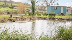 A rustic cabin by a pond with a wooden dock, surrounded by greenery and hills. - Gwithian Shepherds Hut (Hayle)