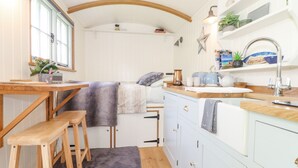 A compact kitchen with wooden stools, a small dining table, and a window with curtains. - Gwithian Shepherds Hut (Hayle)