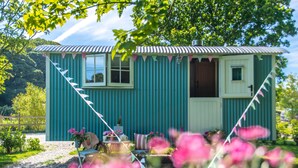 A small, single-story building with a corrugated metal roof and a door. There is a small porch area with a table and chairs. - Gwithian Shepherds Hut (Hayle)