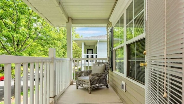 A covered porch with a wicker chair and a small table, a white railing, and a view of a house in the background.