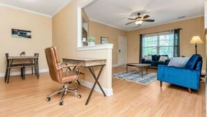 A modern living room with a blue sofa, a brown office chair, a dining table, and a ceiling fan.