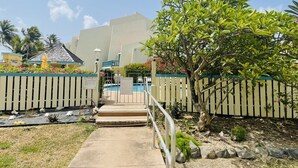 Un camino conduce a una zona de piscina con cerramiento, un pequeño jardín y un edificio al fondo.
