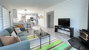 A modern living room with a sofa, coffee table, and TV. There is a kitchen in the background with white cabinets and a microwave. - Encanto's House – A Forest Haven (Marysville)