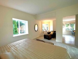A spacious bedroom with a large bed, a sitting area with a chair and table, and a view of greenery through the window.