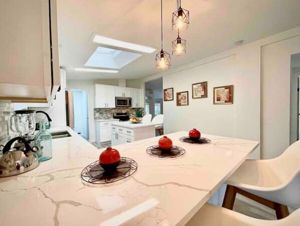 A modern kitchen with white cabinets, a central island with a marble countertop, and a dining area with white chairs.