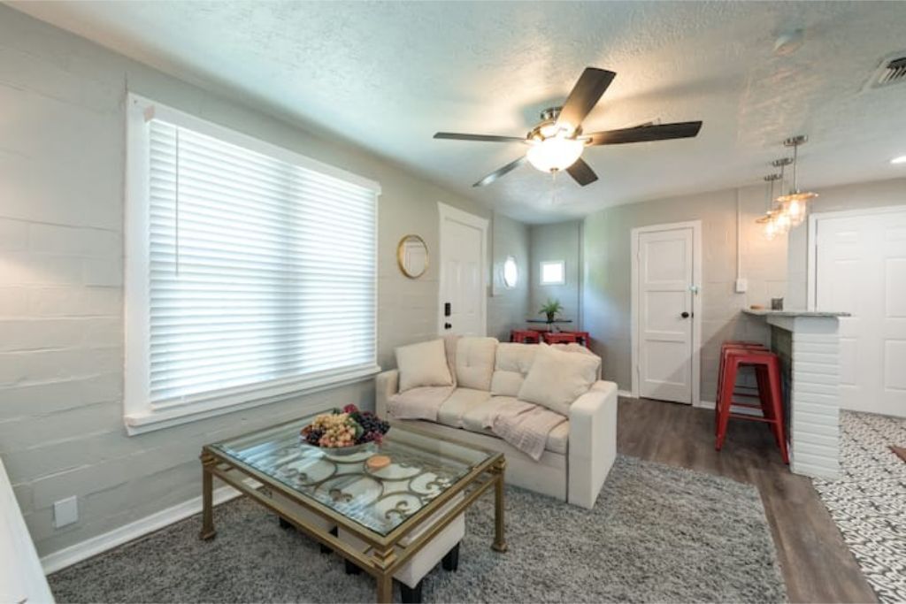 A living room with a glass coffee table, a beige sofa, a ceiling fan, and a kitchen area with a red stool.