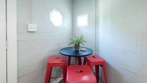 Een kleine kamer met een ronde tafel, twee rode krukken, een bloempot en een klein raam.