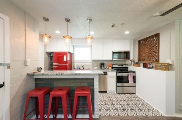 A compact kitchen with white cabinets, a red refrigerator, a microwave, and a stove. There are three red bar stools and pendant lights above the counter.